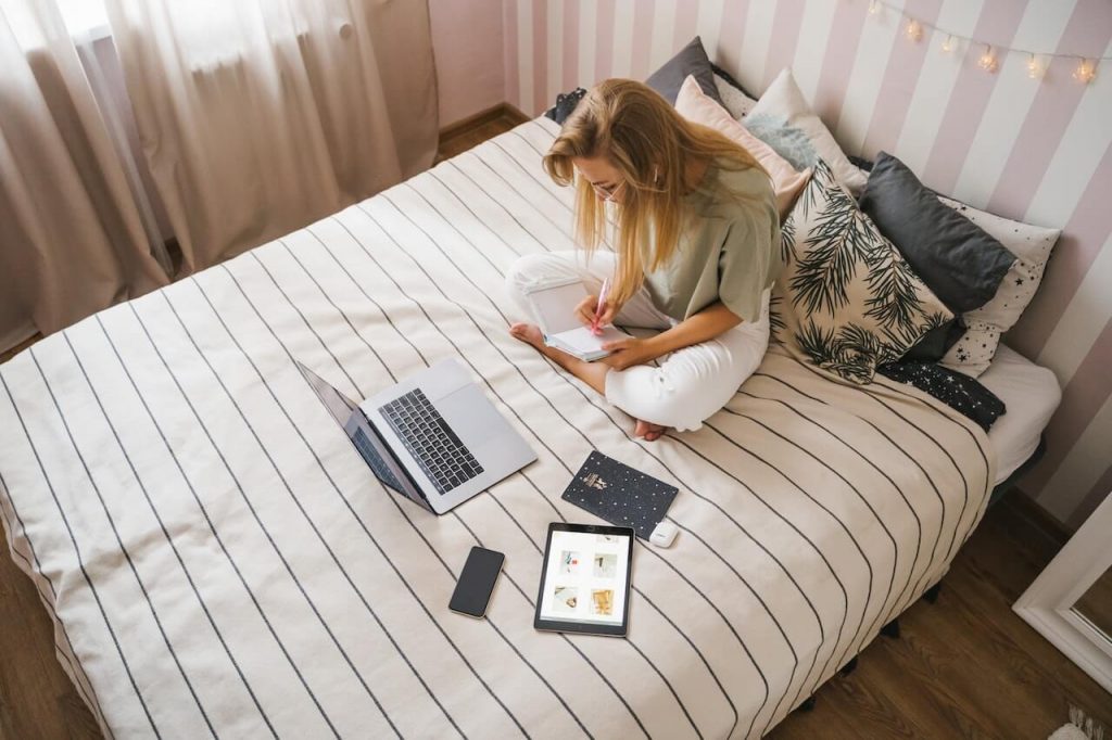 A girl sitting on a bed, writing in a small notebook, with a laptop, tablet, phone, and earpods nearby.