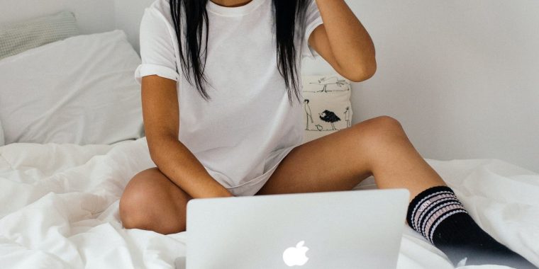A woman on a bed using a laptop, wearing only a t-shirt and knee socks.